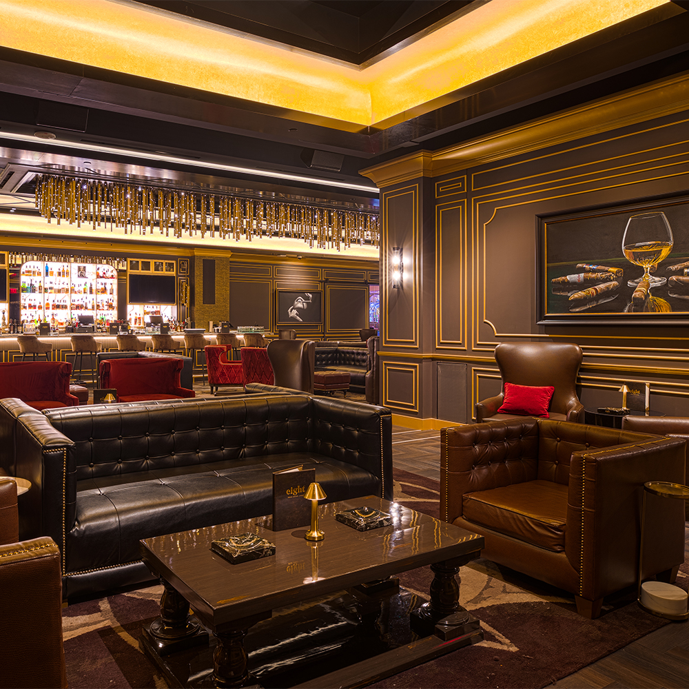 Luxurious cigar lounge with dark leather seating, golden accent lighting, ornate paneling, and a bar with hanging decorative elements. A painting of cigars and a glass of whiskey hangs on the wall.